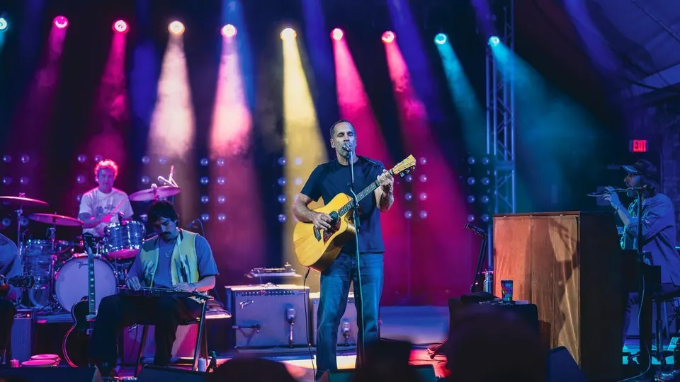 Jack Johnson canta y toca la guitarra acústica en el centro del escenario, con su banda a los lados y focos de colores rosa, amarillo y azul sobre ellos
