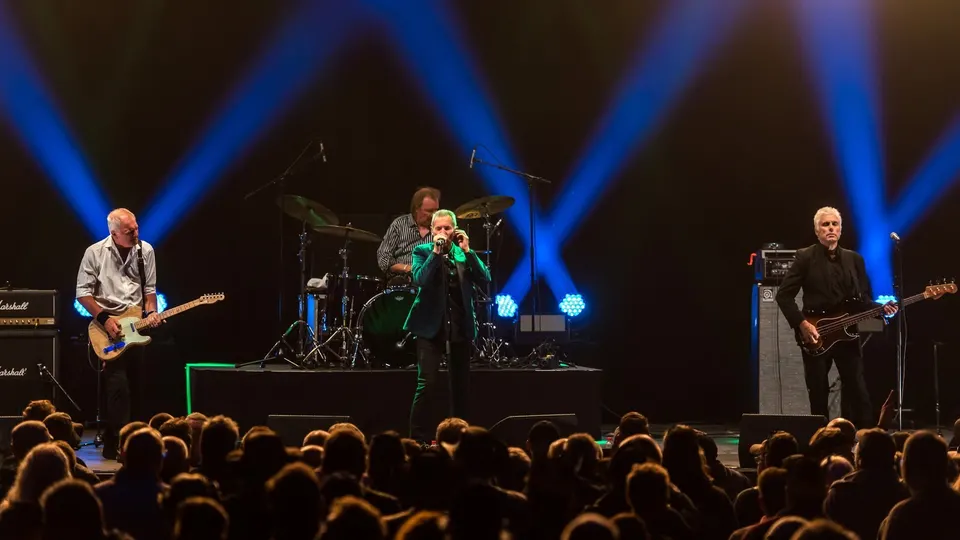 Dr. Feelgood en directo bajo focos azules: el cantante toca la armónica en el centro del escenario, con el guitarrista a la izquierda, el bajista a la derecha y el batería detrás, ante un público a contraluz