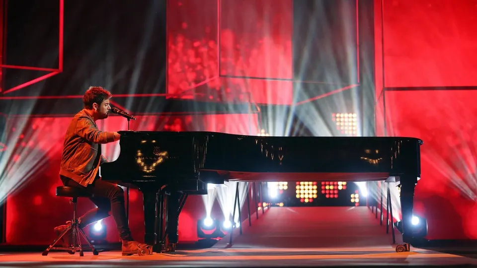 Pablo López canta sentado a un piano de cola negro en un escenario iluminado en rojo, con focos blancos al fondo, durante la gala de los Premios Ondas 2016