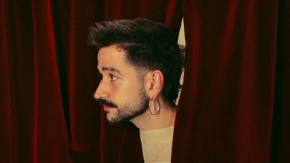 Camilo, de perfil, con bigote, barba y un aro plateado en la oreja, asoma entre unas cortinas de terciopelo rojo