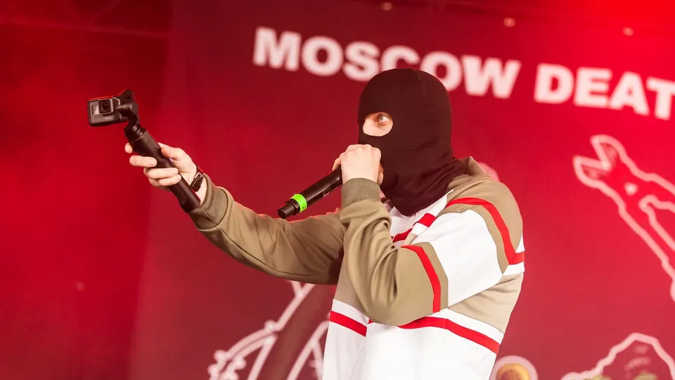 Un integrante de Moscow Death Brigade, con pasamontañas negro y chaqueta de chándal, rapea con el micrófono pegado a la boca y una cámara en la otra mano, delante de una lona roja con el nombre del grupo