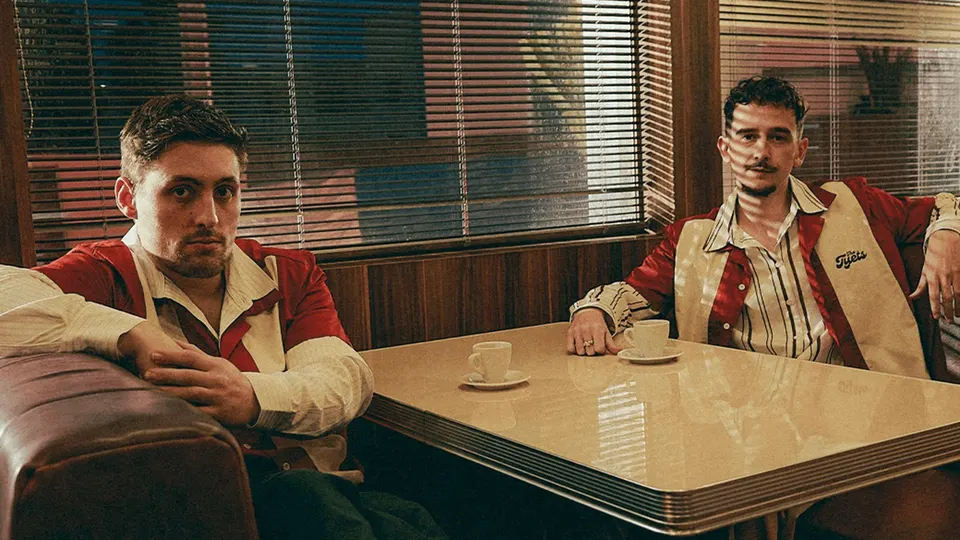 Los dos integrantes de The Tyets sentados a una mesa de cafetería de estilo americano, con chaquetas rojas y blancas, tazas de café y persianas detrás