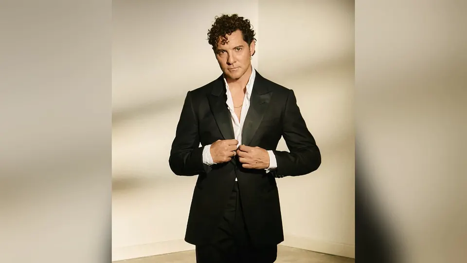 David Bisbal con traje negro y camisa blanca abierta, de pie frente a una pared clara, en la foto promocional del Tour Eternos que publica el Concert Music Festival 2027