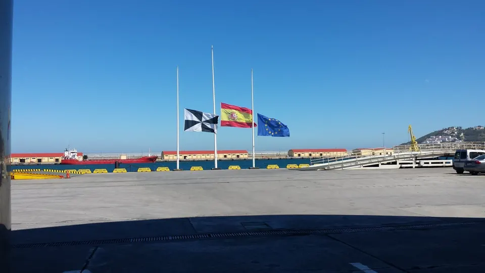 Banderas de Ceuta, España y la Unión Europea a media asta en el puerto de Ceuta, bajo un cielo despejado