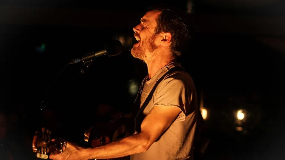 Damien Rice canta con los ojos cerrados ante un micrófono mientras toca la guitarra acústica, iluminado con luz cálida sobre fondo oscuro