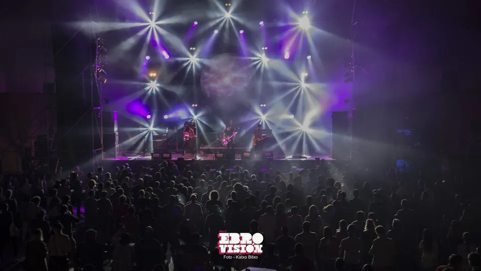 Público de Ebrovisión de espaldas frente al escenario principal, con una banda tocando bajo haces de luz blanca y morada y humo