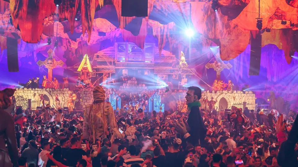 Pista de una fiesta Horroween de elrow llena de público, con un decorado de castillo gótico, cruces con calaveras, telas rojas y naranjas colgadas del techo y muñecos gigantes de una momia y Frankenstein entre la gente