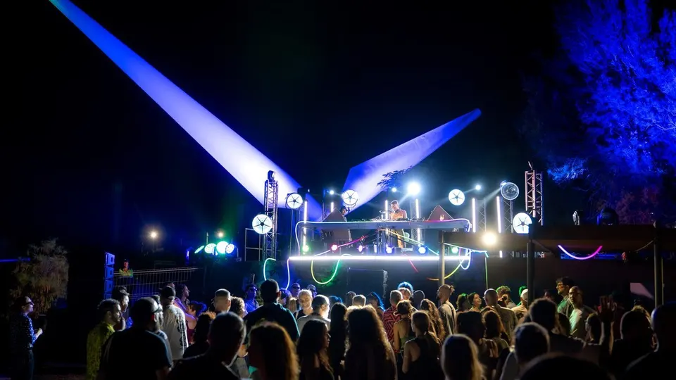 Público frente a una cabina de DJ al aire libre en el ITER de Tenerife, de noche, con dos grandes aspas de aerogenerador iluminadas en azul sobre el escenario de Eólica