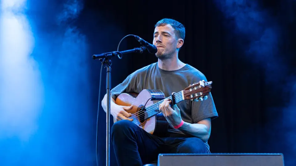 Charlie Cunningham, sentado con una guitarra acústica, canta ante un micrófono en un escenario iluminado de azul