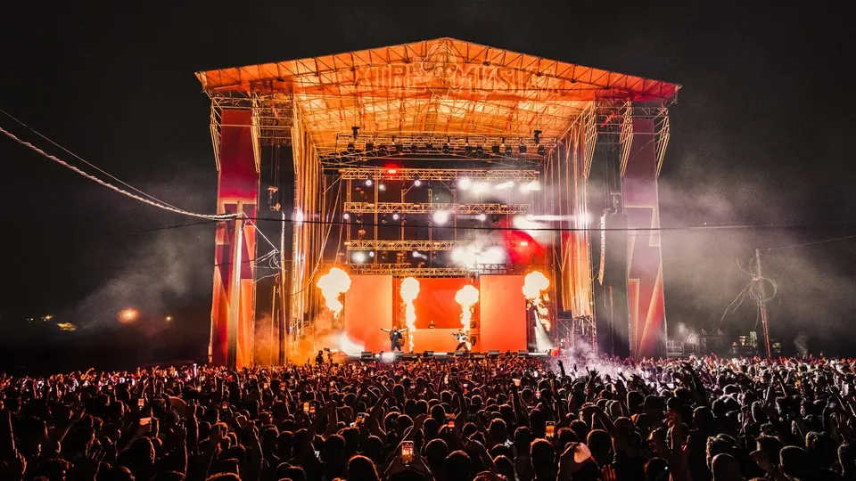 Escenario principal de Extremúsika de noche, con llamaradas de pirotecnia, luces naranjas y dos artistas sobre el escenario ante miles de personas con los brazos en alto