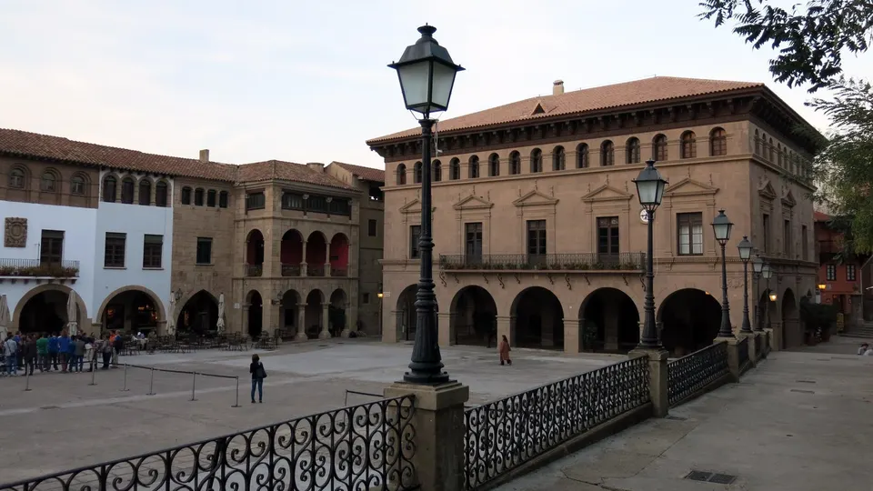 La plaça Major del Poble Espanyol de Barcelona al atardecer, casi vacía, con la fachada porticada de la casa consistorial de Vall-de-roures, farolas de hierro y una barandilla en primer plano