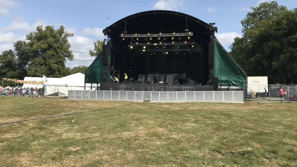 Escenario de un festival al aire libre con la explanada de césped vacía delante, las vallas montadas y las luces encendidas antes de la llegada del público