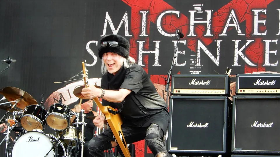 Michael Schenker, con gorro de pelo y gafas de sol sobre la frente, sonríe mientras toca una guitarra amarilla en forma de V delante de su nombre en la pantalla y de una pared de amplificadores Marshall