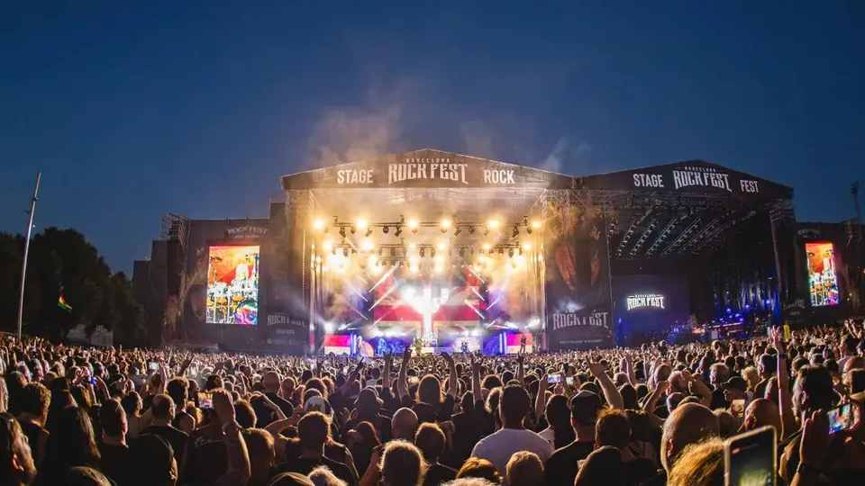 Escenario principal de Barcelona Rock Fest de noche, con las lonas del festival iluminadas, pantallas a los lados y miles de personas con los brazos en alto frente al escenario