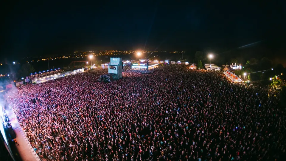 Vista aérea nocturna del recinto del FIB en Benicàssim lleno de público frente al escenario principal, con la torre de control rotulada FIB en el centro y las luces del pueblo al fondo
