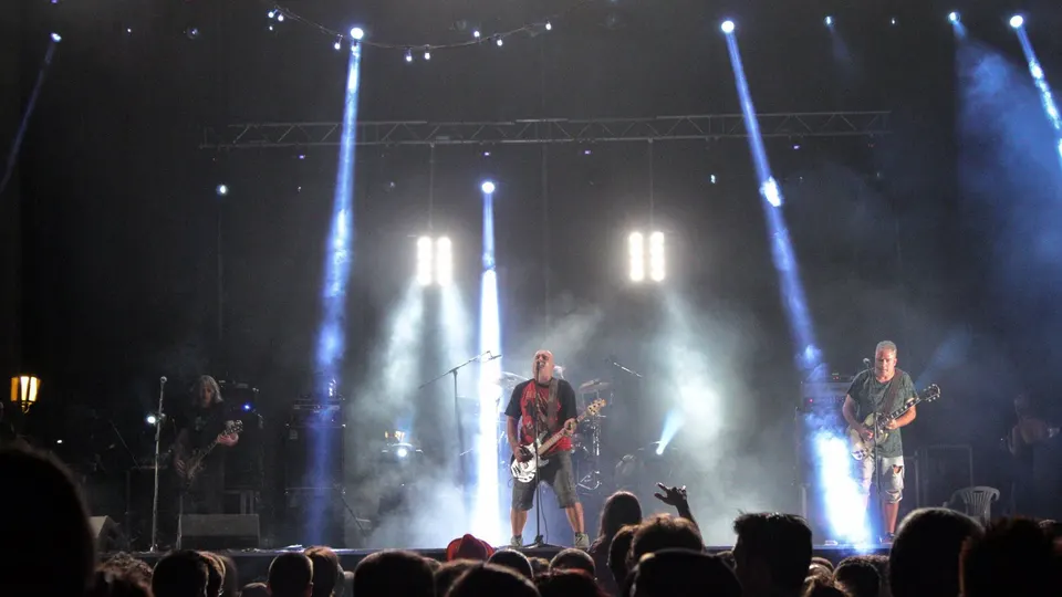 Reincidentes tocan en un escenario al aire libre bajo haces de luz blanca y azul y humo: el bajista y cantante en el centro, con camiseta roja, y dos guitarristas a los lados, con el público de espaldas en primer plano