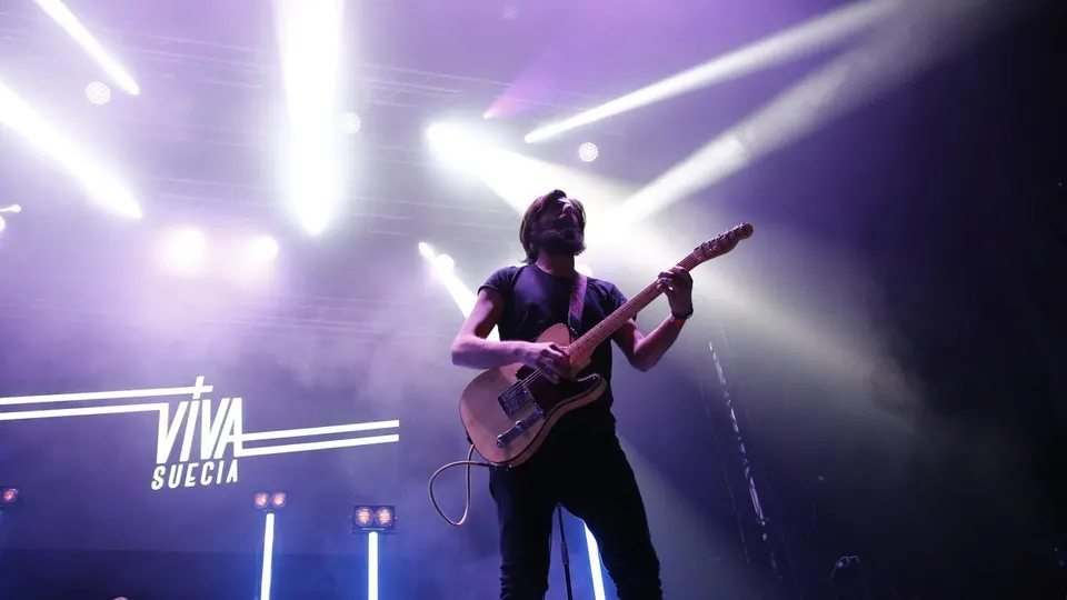 Un guitarrista de Viva Suecia toca una guitarra eléctrica bajo focos violetas, con el logotipo del grupo iluminado a su derecha en el escenario de Palencia Sonora