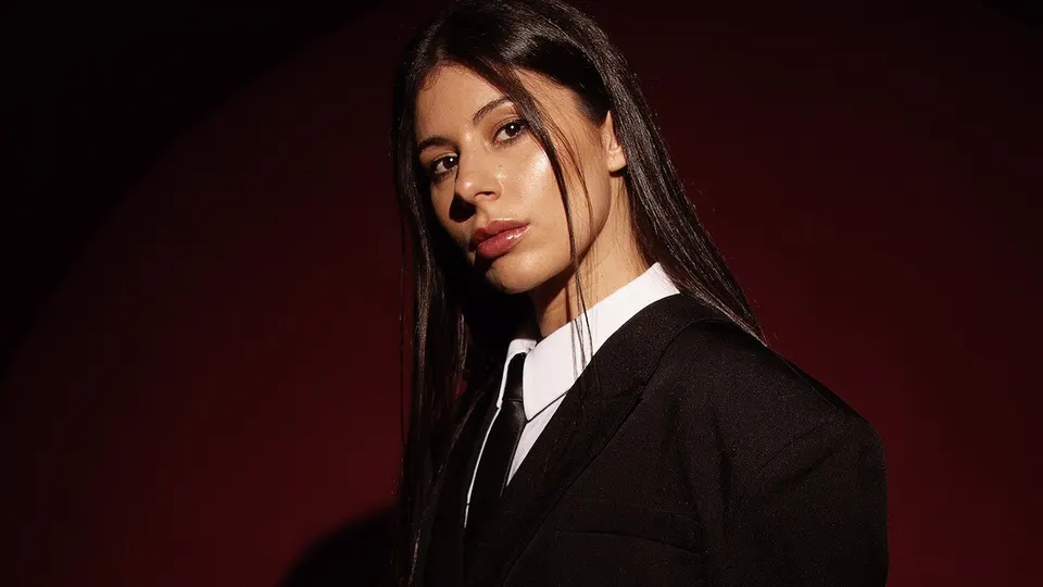 Retrato de la DJ Bárbara Lago, con el pelo largo y oscuro, traje negro, camisa blanca y corbata, sobre un fondo rojo oscuro