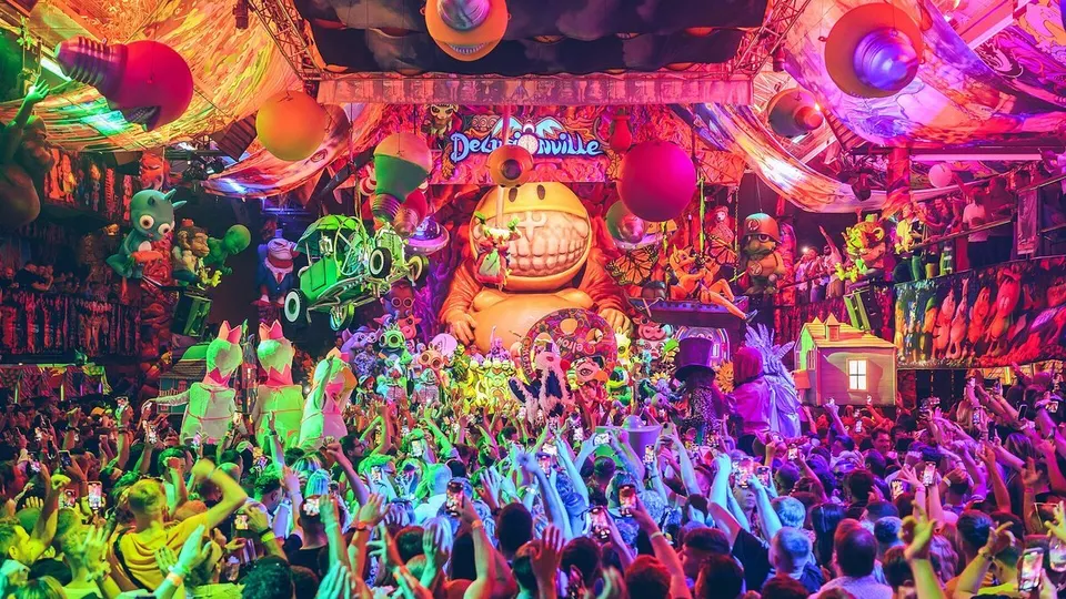 Público con los brazos en alto ante una escenografía de elrow llena de muñecos gigantes, globos y luces de colores alrededor de la cabina del DJ
