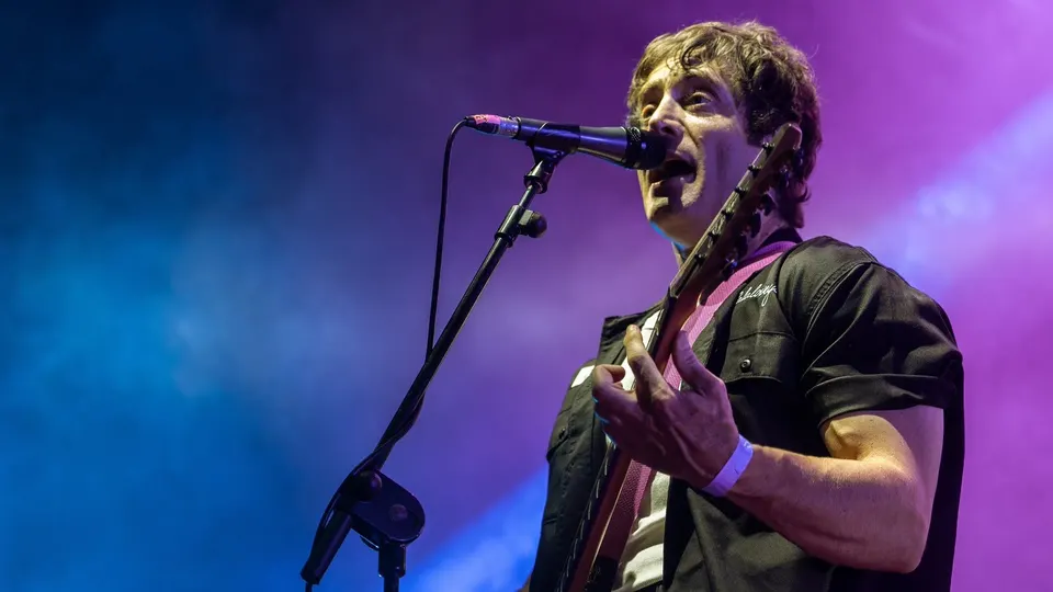 El cantante y guitarrista de La La Love You canta ante el micrófono con camisa negra, bajo luces azules y violetas