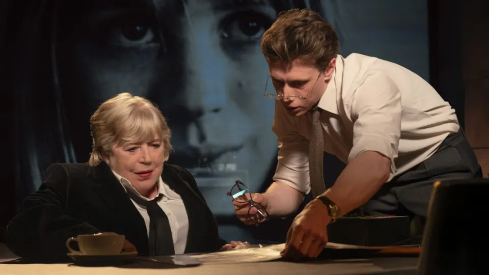 Fotograma de Broken English: Marianne Faithfull, sentada ante una mesa con una taza de café, mientras un actor con camisa y corbata se inclina sobre unos papeles con unas gafas en la mano; al fondo, un gran retrato proyectado de un rostro joven
