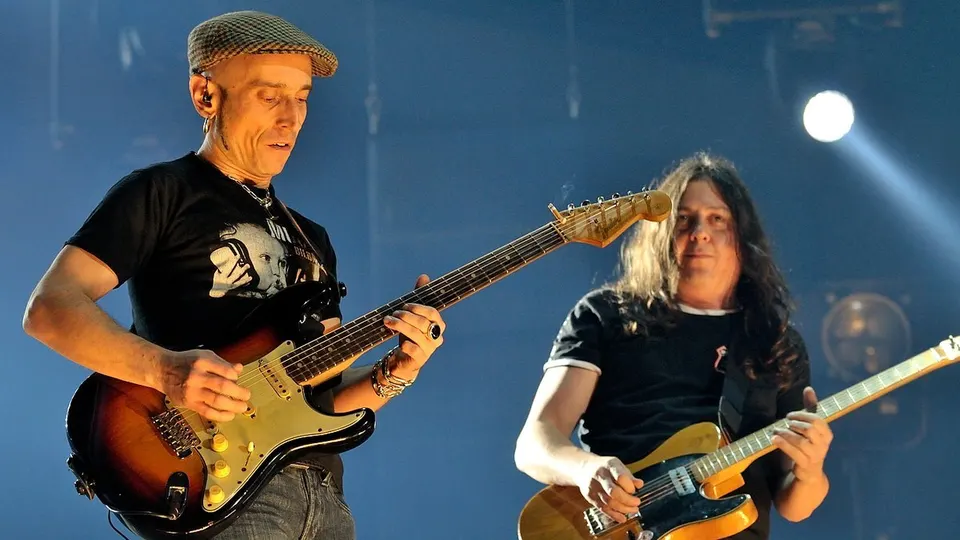Fito Cabrales, con gorra de cuadros y camiseta negra, toca una guitarra eléctrica junto a otro guitarrista de Fito & Fitipaldis sobre un escenario iluminado en azul