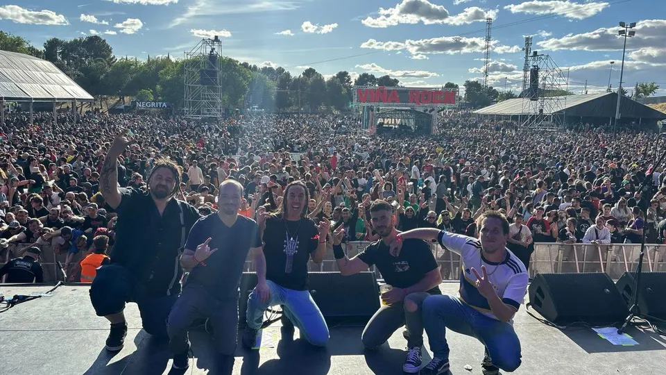 Los músicos de Kaos Etíliko posan agachados en el borde del escenario de Viña Rock 2024, con miles de personas detrás bajo un cielo azul con nubes