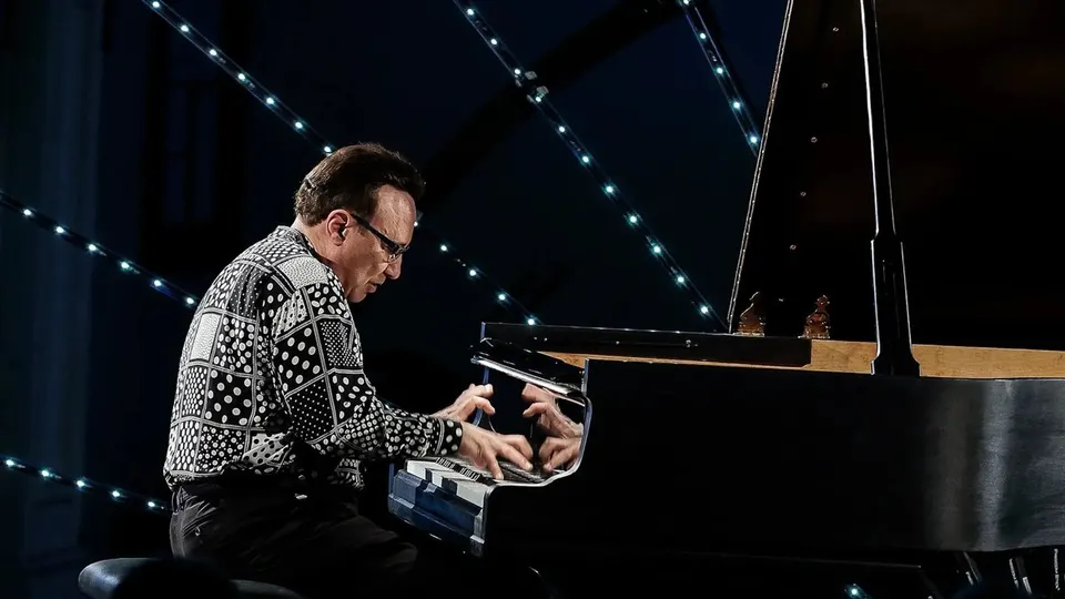 Michel Camilo, con gafas y camisa blanca y negra de lunares, toca un piano de cola negro en un escenario oscuro cruzado por hileras de luces