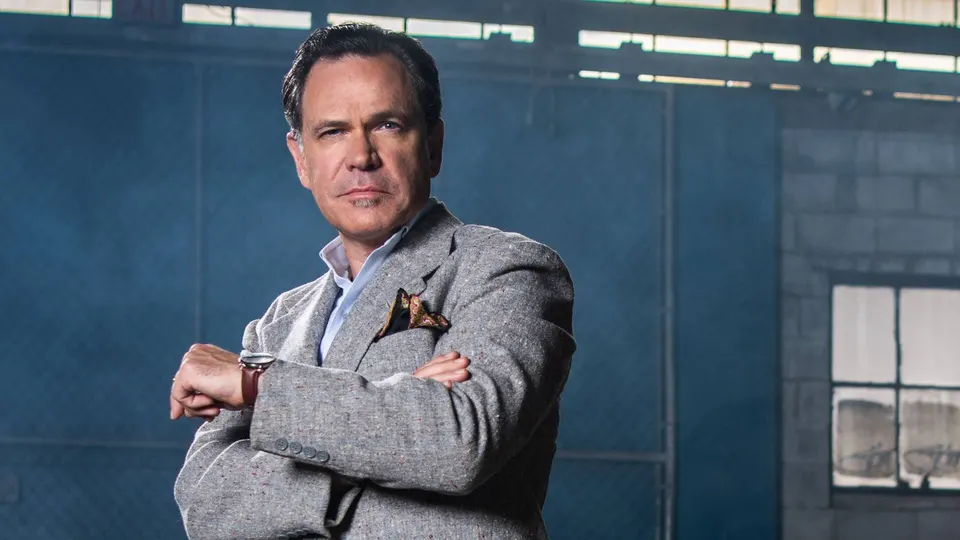 Retrato del cantante Kurt Elling con americana gris de tweed y los brazos cruzados en una nave industrial con paredes azules y ventanales, en una foto de promoción