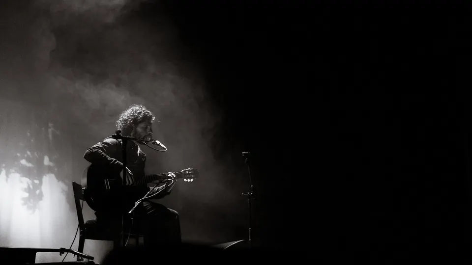 José González, sentado en una silla, canta y toca la guitarra española en un escenario a oscuras, iluminado de lado entre humo, en una foto en blanco y negro