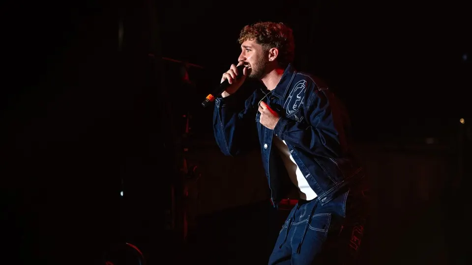 Lucho RK canta con el micrófono en la mano, vestido con chaqueta y pantalón vaquero, sobre un escenario a oscuras