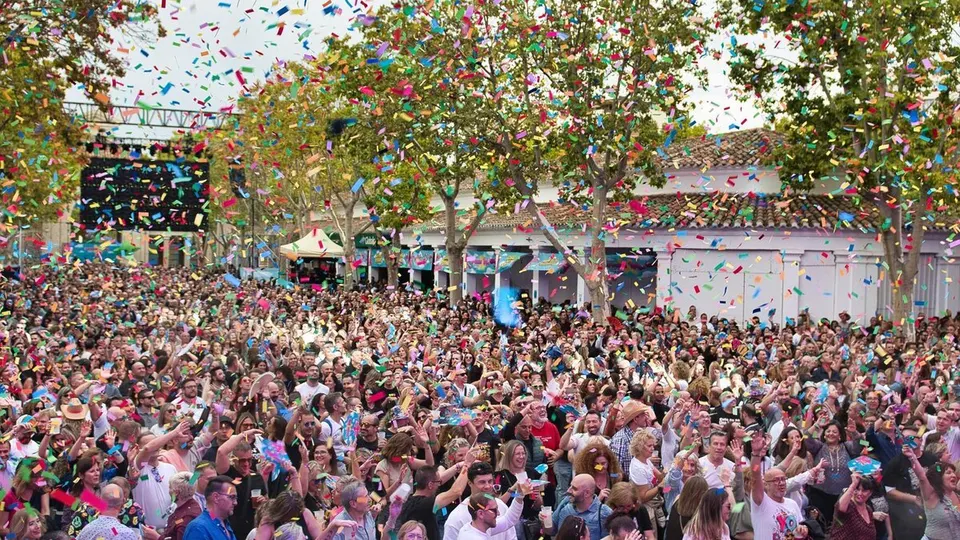 Una multitud con los brazos en alto bajo una lluvia de confeti de colores en el Recinto Ferial de Albacete, con los árboles y los soportales blancos del recinto al fondo, durante Lunátikko 90s