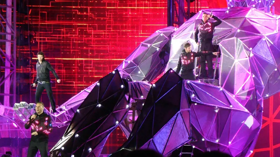 Take That actúan con chaquetas de luces sobre una gran estructura de cristales violetas, con una pared de luces rojas al fondo, en la gira Progress Live de 2011