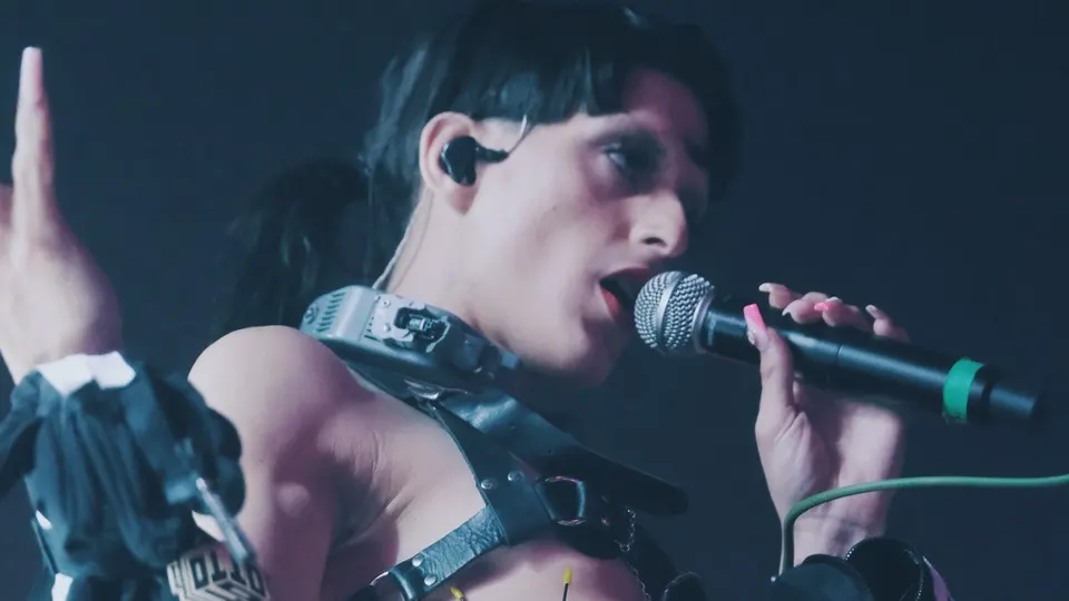 Arca canta con un micrófono en la mano y un arnés de cuero, con el pelo negro recogido y un auricular de escenario, sobre fondo oscuro en el Sónar de Barcelona