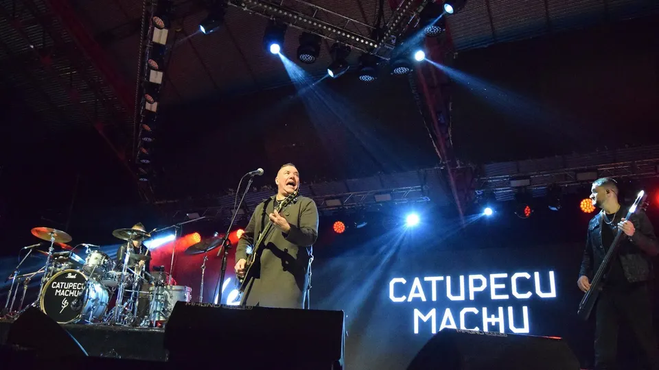 Fernando Ruiz Díaz canta y toca la guitarra con abrigo verde al frente de Catupecu Machu, con el batería detrás, un bajista a la derecha y el nombre del grupo en la pantalla del escenario bajo focos azules