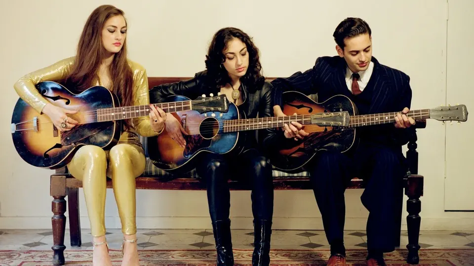 Kitty, Daisy & Lewis sentados en un banco de madera tocando tres guitarras acústicas antiguas: una mujer con mono dorado, otra con chaqueta de cuero negro y un hombre con traje de raya diplomática