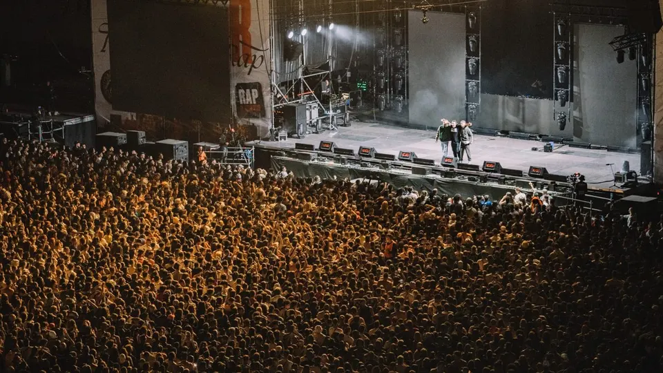 Miles de personas llenan la explanada ante el escenario principal de Rocanrola mientras tres artistas saludan al público al final de un concierto