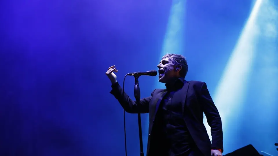 Iván Ferreiro canta con el brazo levantado frente a un micrófono de pie, vestido de negro y bajo focos azules, en un escenario de festival