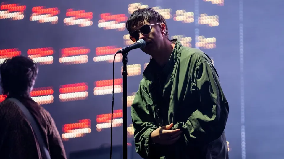 Julian Casablancas, cantante de The Strokes, con gafas de sol y chaqueta verde ante el micrófono, con luces rojas en la pantalla del fondo