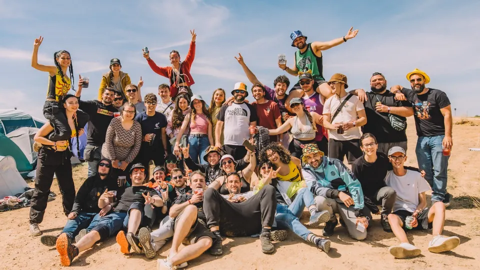 Una treintena de asistentes de Viña Rock posan sonrientes en la zona de acampada, sobre tierra y bajo un cielo azul, con tiendas de campaña al fondo, gorros, gafas de sol y brazos en alto