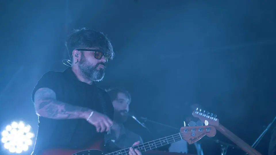 Un músico de Viva Suecia, con gafas y barba, toca el bajo iluminado de azul en el escenario de Brisa Festival, con otro músico detrás
