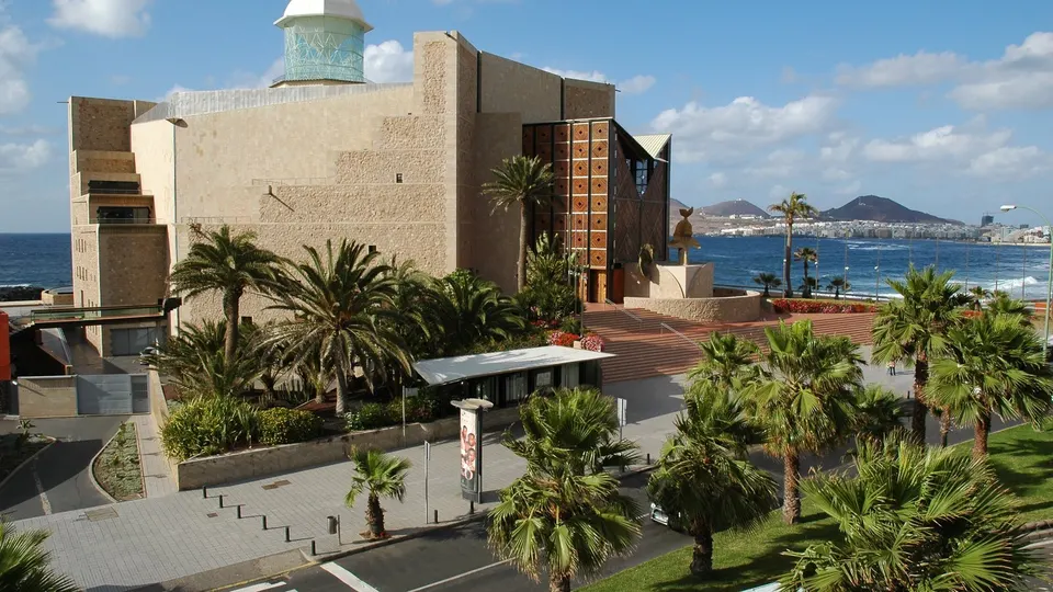 Auditorio Alfredo Kraus de Las Palmas de Gran Canaria, con su fachada de piedra, palmeras delante y el mar y la playa de Las Canteras al fondo