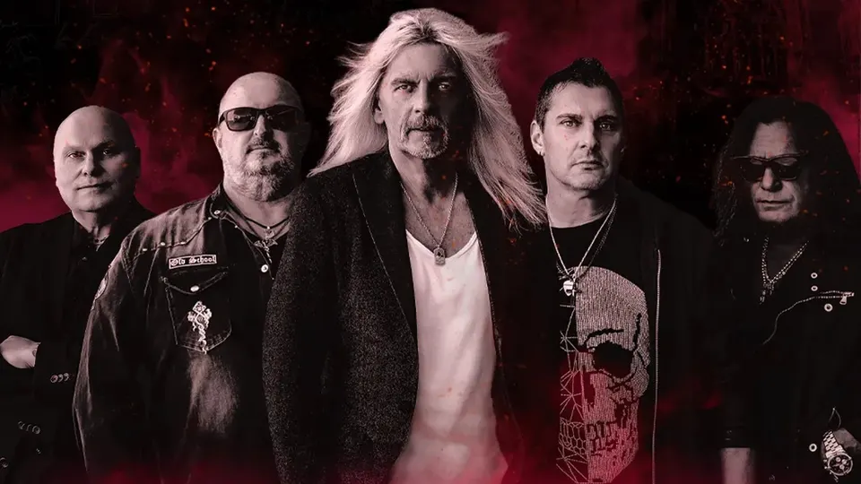 Axel Rudi Pell, con el pelo largo y rubio y camiseta blanca, en el centro de su banda de cinco músicos vestidos de negro, sobre un fondo rojo oscuro con chispas