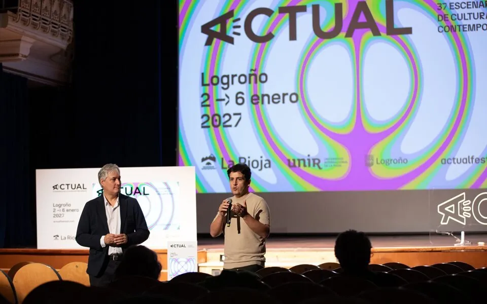 Vista desde el patio de butacas de la presentación de Actual 2027: dos hombres en el escenario ante la pantalla con la figura de vibración del festival, los logotipos de La Rioja, UNIR y Logroño y un atril con el cartel impreso