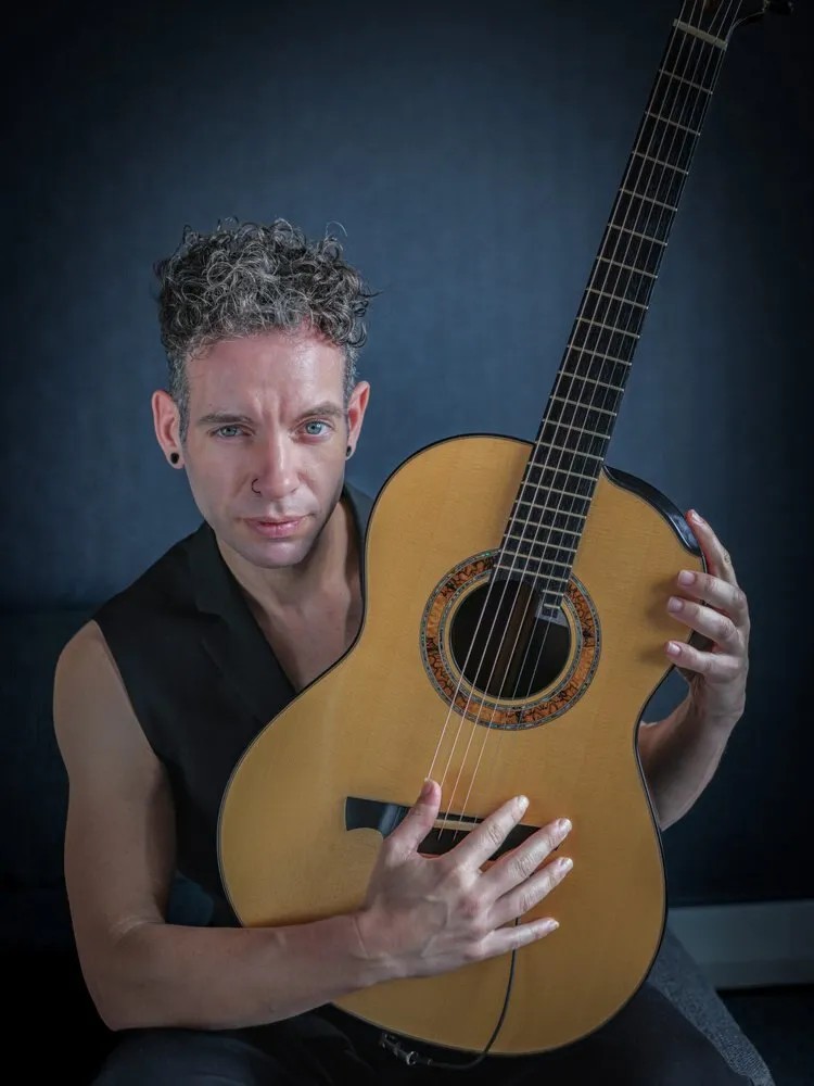 Guitarist Pipo Romero, with curly grey hair and a black sleeveless T-shirt, holds an acoustic guitar against a dark blue background
