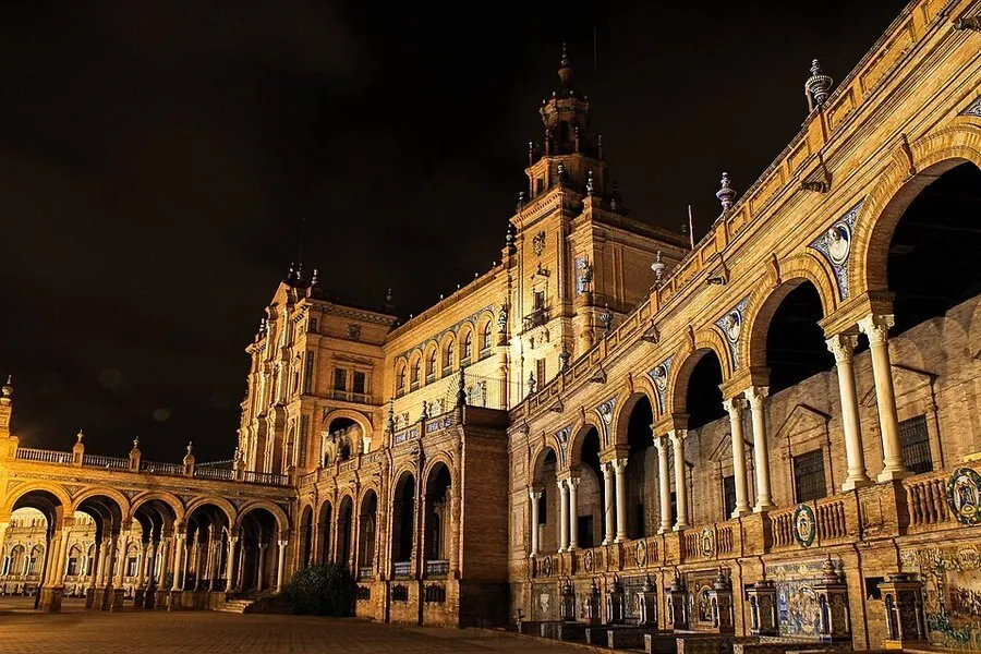 La Plaza de España de Sevilla de noche, con su arquería y su torre iluminadas en tonos dorados bajo el cielo oscuro