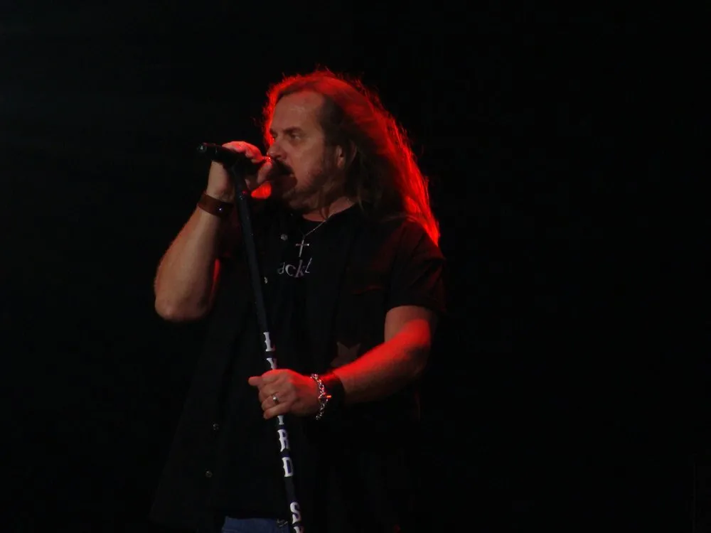 Johnny Van Zant, cantante de Lynyrd Skynyrd, canta agarrado al pie de micrófono con el nombre del grupo, iluminado en rojo sobre un fondo negro