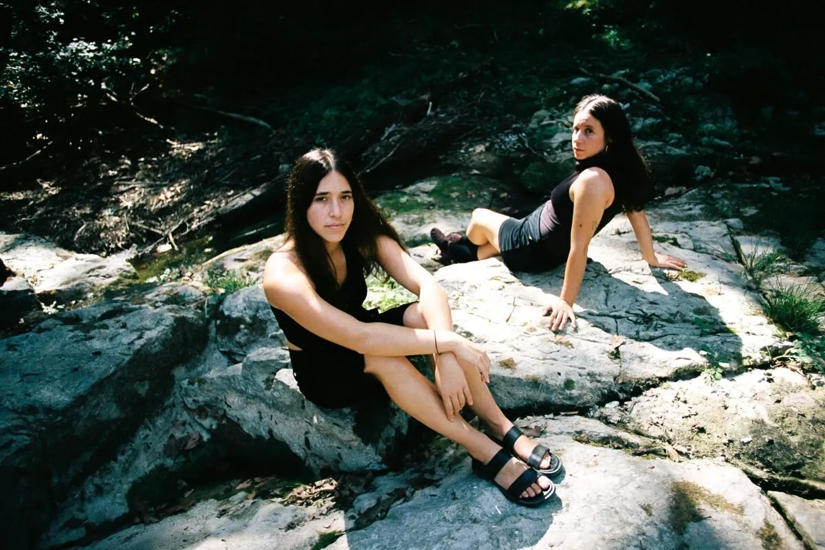 Dos mujeres jóvenes de pelo largo y oscuro, vestidas de negro, sentadas sobre grandes rocas claras en un bosque soleado; una mira a cámara en primer plano y la otra, detrás, se apoya con las manos sobre la piedra