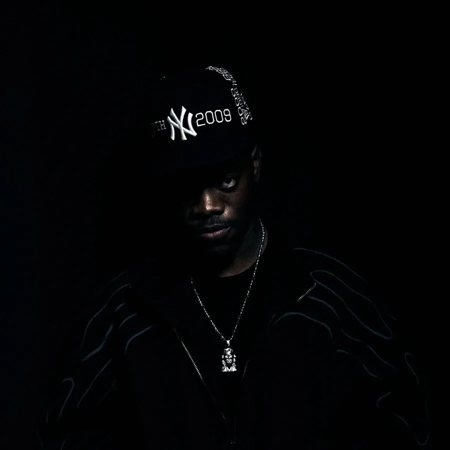 Retrato en penumbra del rapero Brocki con gorra negra de los New York Yankees, chaqueta oscura y una cadena plateada, con media cara iluminada