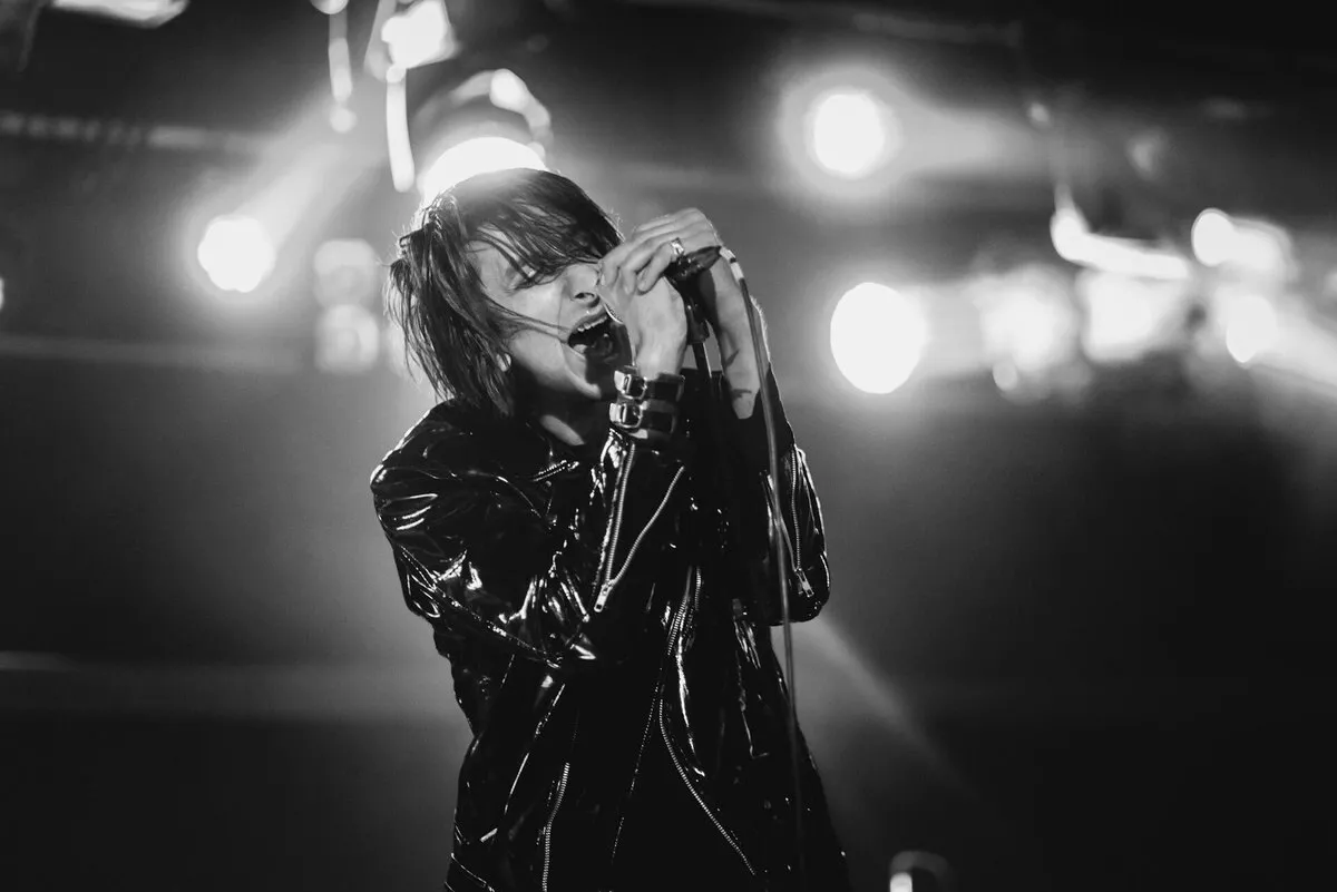 Faris Badwan, cantante de The Horrors, canta con las dos manos sobre el micrófono, con chaqueta de cuero y focos detrás, en una foto en blanco y negro